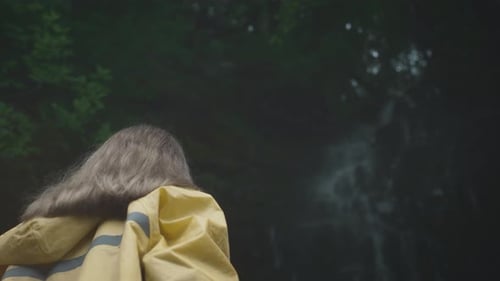 Caucasian Girl Traveler in a Yellow Raincoat Makes a Photo of a Waterfall on a Smartphone in the