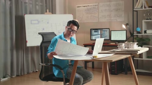 Asian Man Engineer Looking At Blueprint And Typing On A Laptop At The Office