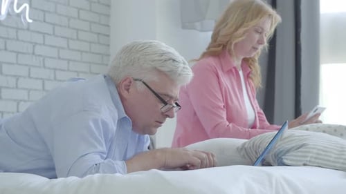 Side View of Senior Caucasian Man Lying on Bed and Using Laptop As Woman with Smartphone Sitting