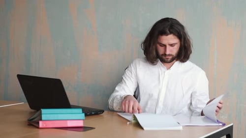 Young Beraded Man with Long Hair in White Shirt Working at Office with Papers and Laptop