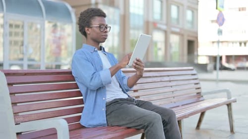 Junger afrikanischer Mann benutzt Tablet beim Sitzen im Freien auf der Bank