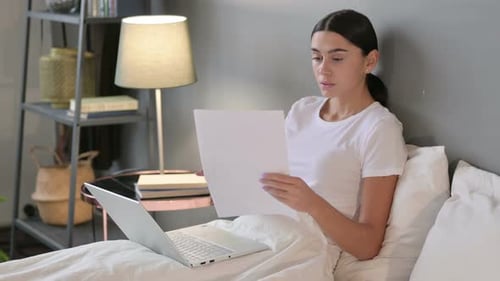 Woman Reviews Documents in Bed with Laptop