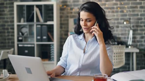 Beautiful Girl Speaking on Mobile Phone Using Laptop and Smiling in Office