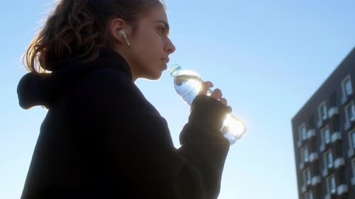Young Woman Sits on Bench in Apartment Complex in Morning After Jogging and Drinks Water