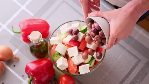 Griechische Salatzubereitungsserie Concept Woman gießt Oliven in Schüssel mit gehacktem Gemüse