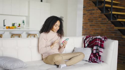 Curly Brunette Girl Holding a Smartphone in Her Hands While Sitting on the Sofa in the Studio