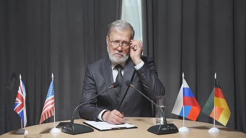 Man at Table with Flags Speaking to Camera
