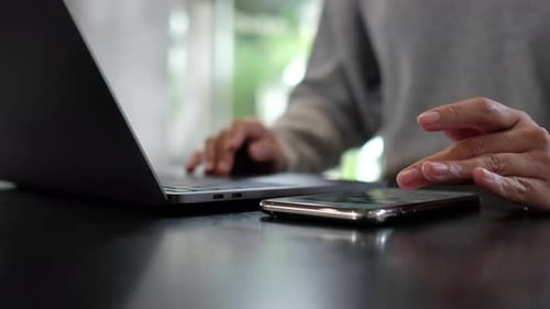 Nahaufnahme einer Frau, die den Smartphone-Bildschirm berührt und scrollt, während sie am Laptop arbeitet
