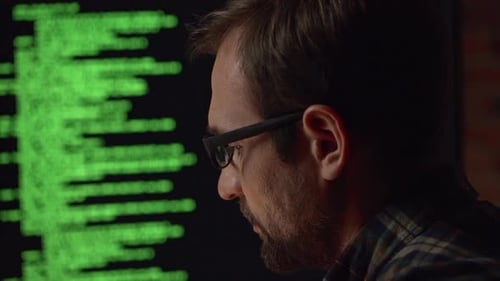 Nahaufnahme eines kaukasischen Mannes mit Brille, der in Zeitlupe auf den Monitor mit grünem Text schaut