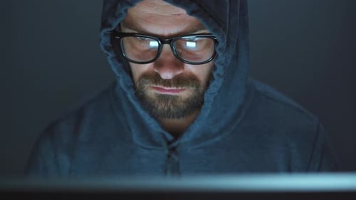 Hacker masculino en el capó y gafas trabajando en una computadora en una sala de oficina oscura