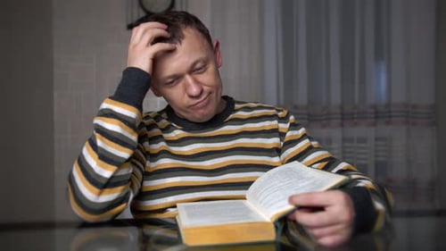 Müder junger Mann studiert den Text in einem Buch, während er am Tisch sitzt