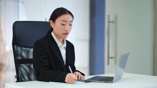 Young Focused Asian Businesswoman Studying Document on Laptop and Writing Notes Working at Her