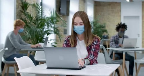 Young Focused Caucasian Woman in Covid-19 Face Mask Messaging Online in Office with Multiethnic