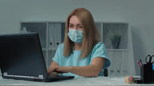 Young Woman Puts a Medical Mask on Her Face and Works at a Laptop in the Office