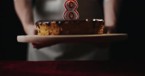 Young Woman Wearing Kitchen Apron Puts Homemade Chocolate Cake or Pie with Number 8 Burning Candle
