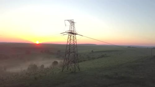 Aerial view of metal power tower with high voltage electric wires at dawn in rural landscape.