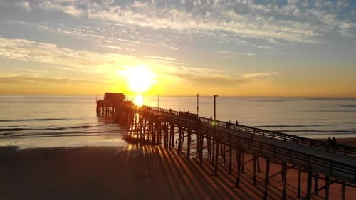 Drone rising over the Newport Beach pier at sunset over the pacific ocean as people walk along the C