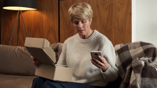 Woman Opens Box While Using Cell Phone Indoors