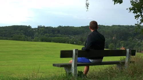 Young Man Sits on the Bench in Nature (Field with Forest) and Looks Around