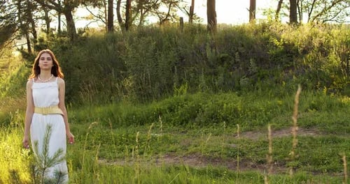 A Cheerful Teenage Girl in Light Dress Walks in a Clearing Between Green Trees in Summer Forest