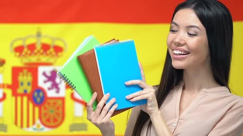 Femme souriante montrant des cahiers à la caméra, drapeau espagnol, fond, étude à l'étranger