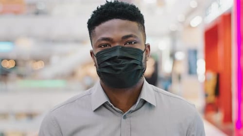 Nahaufnahme Junger afroamerikanischer Mann in medizinischer Schutzmaske, der im Einkaufszentrum spaziert und in die Kamera schaut