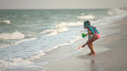 A Girl with African Braids in a Summer Costume Plays on the Beach with Shells Near the Sea with