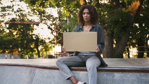 Afroamerican Woman Showing Empty Cardboard Poster Posing Sitting on Pump Track at Rollerdrome in