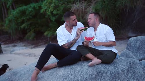 Feliz casal homossexual caucasiano comendo melancia sentado em uma rocha na ilha tropical ao entardecer