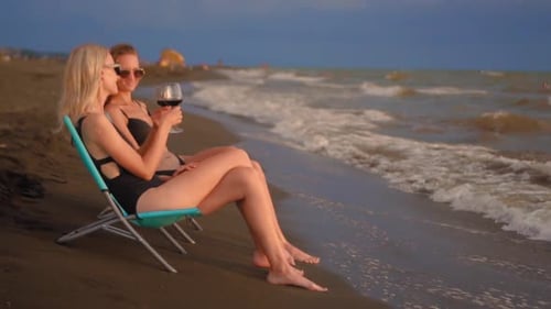 Two women sitting by sea or ocean and drinking red wine from glasses.