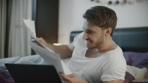 Excited Man Reviews Documents in Bed with Laptop