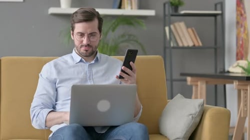Man Works on Laptop and Phone on Couch