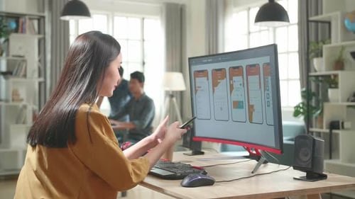 Asian Female Mobile Application Developer Works And Using Mobile Phone With Graphics On Computer
