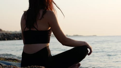 A yogi woman sits in a lotus position on the seashore and looks at the sea dawn of the sun