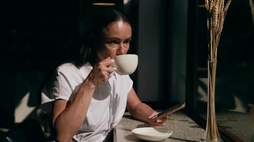 Middle Aged Woman is Enjoying Cup of Coffee in Cafe at Sunny Day and Surfing Internet By Cellphone