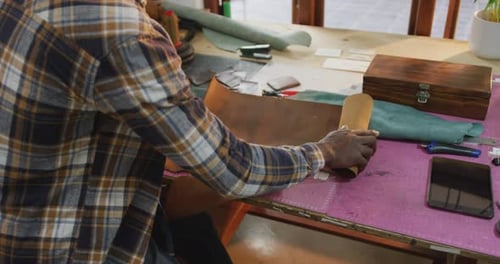 Midsection of african american craftsman unrolling leather in leather workshop