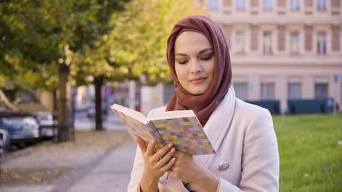 A Young Beautiful Muslim Woman Reads a Book with a Smile in a Street in an Urban Area Closeup