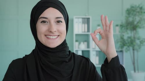 Portrait Indoors Muslimisches islamisches Mädchen lächelt zahnweit in die Kamera und zeigt Hand Ok Schild
