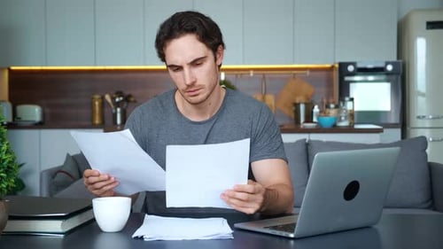 Young Employee Works at Laptop Remotely Analyzes Graph on Paper and Verifies the Content with the