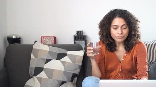 Woman drinking water and using laptop at home