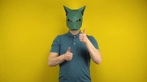 Ein junger Mann in einer Pappschakal-Maske zeigt seinen Daumen hoch auf gelbem Hintergrund. Mann in einem Grün