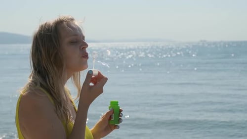 Blonde junge Frau bläst Blasen auf dem Hintergrund des Blauen Meeres. Sie genießt das und ist glücklich.