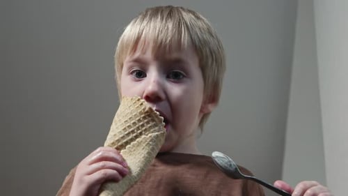 Down View of Little Boy Sits on Table and Eats Icecream with Spoon in Kitchen at Home