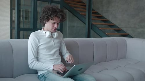 Relaxed Serious Millennial Guy Student Freelancer Using Laptop Device Leaning on Sofa at Home Office