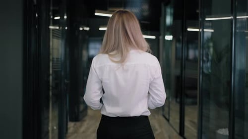 The Camera Follows the Back of a Blonde Business Woman in a Shirt Down the Corridor