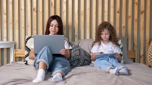 Young Mother Works on Laptop and Daughter Swipes on Tablet