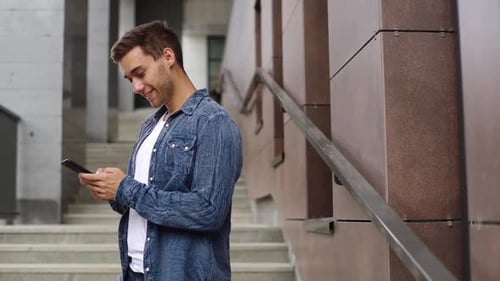 Side View of Handsome Young Man Looking on Smartphone Screen and Laughing Standing on Stairs of