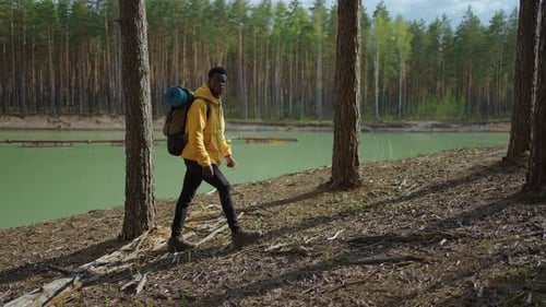 A Young Black Man in a Yellow Cursor Climbs Up with a Backpack Uphill in the Woods Looking at the