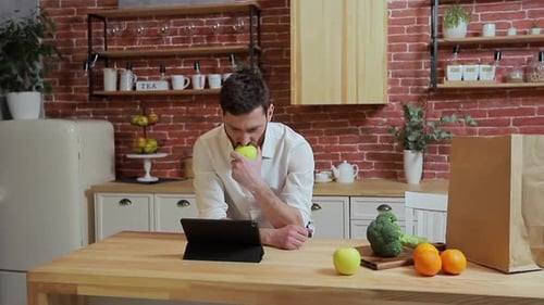 Man Eating Apple While Using Tablet in Kitchen