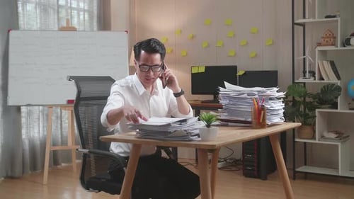 Asian Man Talking On Phone And Taking Note While Working Hard With Documents At Office